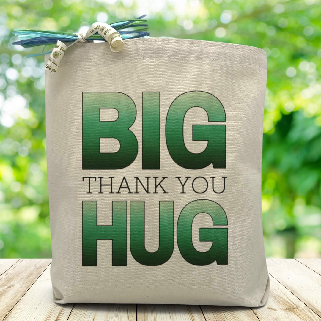 Green Big Thank You Hug printed on a canvas tote bag sitting on a wood table with blurred green background.