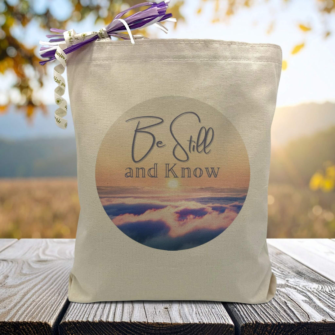 Be Still and know text with sunset background on a canvas tote bag, sitting on wood table with mountains in background.