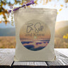 Be Still and know text with sunset background on a canvas tote bag, sitting on wood table with mountains in background.