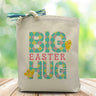 A pink, blue and yellow plaided big easter hug text on a canvas tote bag placed on a wood table with blurred green background.