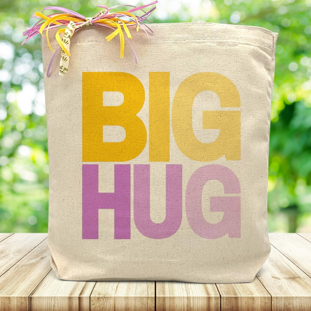 A purple and yellow spring inspired text on a canvas tote bag placed on a wood table with green blurred background.