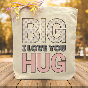 A pink and purple heart filled big i love you text on a canvas tote bag placed on a wood table with tree background.
