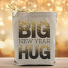 A sparkly big new year hug text on a canvas tote bag placed on a wood table with a light filled background.