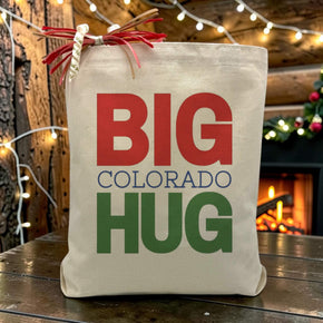 Big Colorado Hug Red and Green Text on a canvas tote bag sitting on a wood table with fireplace in background.