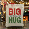 Big Colorado Hug Red and Green Text on a canvas tote bag sitting on a wood table with fireplace in background.