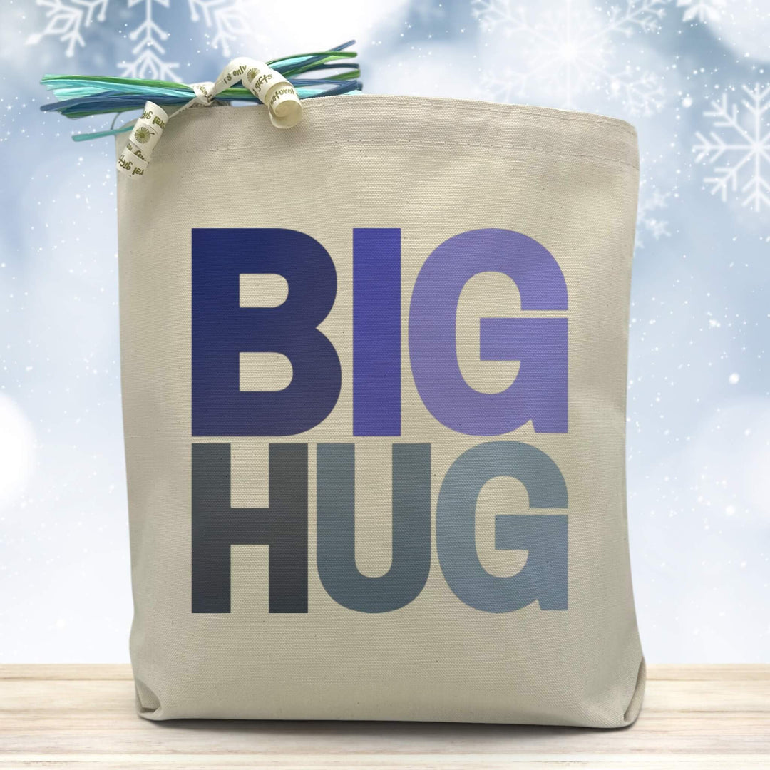 A variation of winter blue colors on Big Hug Text placed on a canvas tote bag sitting on a wood table with snowflakes in the background 