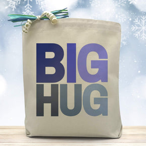 A variation of winter blue colors on Big Hug Text placed on a canvas tote bag sitting on a wood table with snowflakes in the background 