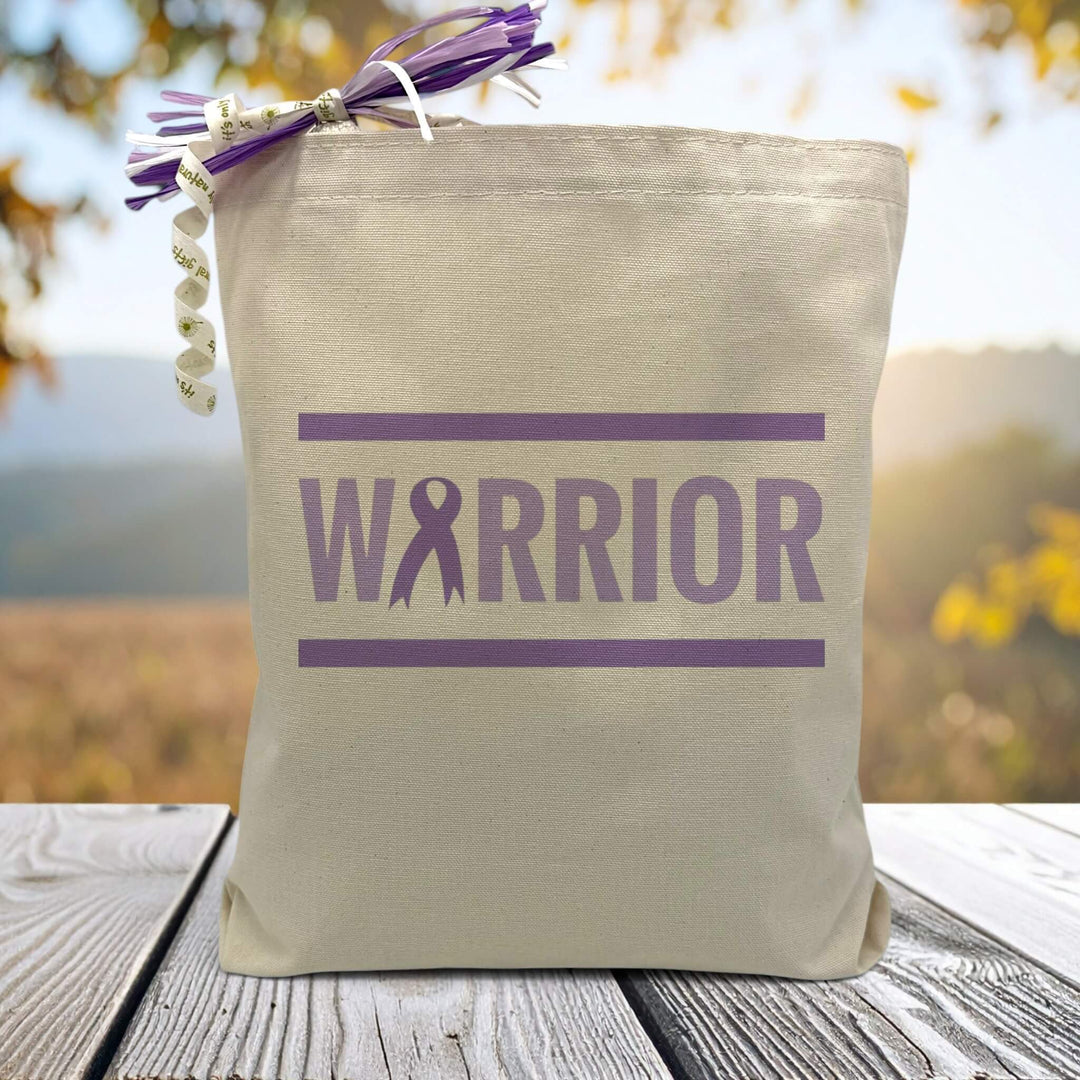 A purple text that says warrior with a cancer ribbon on a canvas tote bag, placed on a wooden table with trees in background.