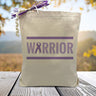 A purple text that says warrior with a cancer ribbon on a canvas tote bag, placed on a wooden table with trees in background.