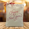 A red cheers to a new year text with stars on a canvas tote bag placed on a wooden table with a light filled background.