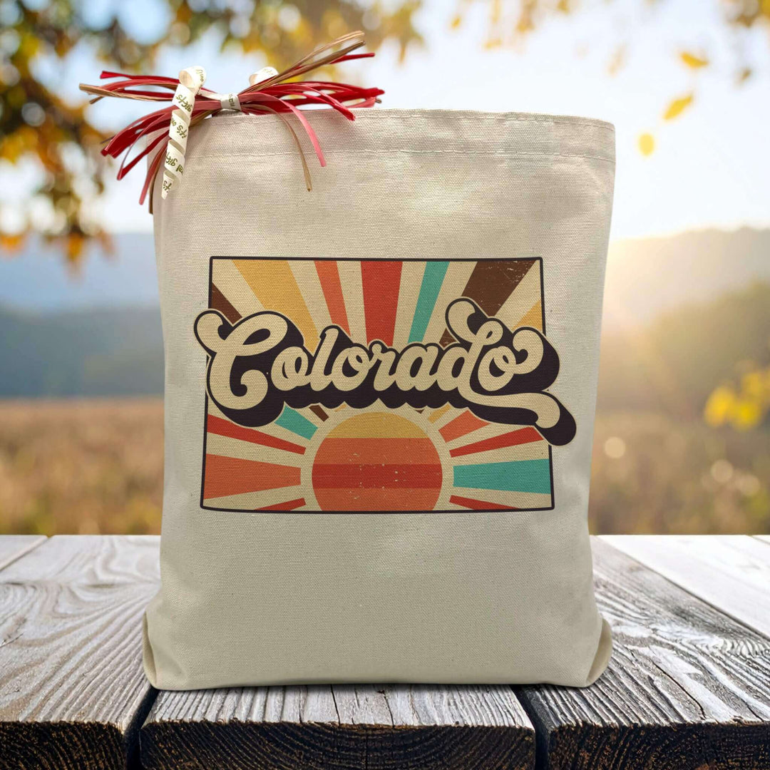 A retro styled rising sun with the word colorado in the middle on a canvas tote bag placed on a wooden table with trees in background.