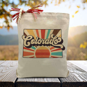 A retro styled rising sun with the word colorado in the middle on a canvas tote bag placed on a wooden table with trees in background.