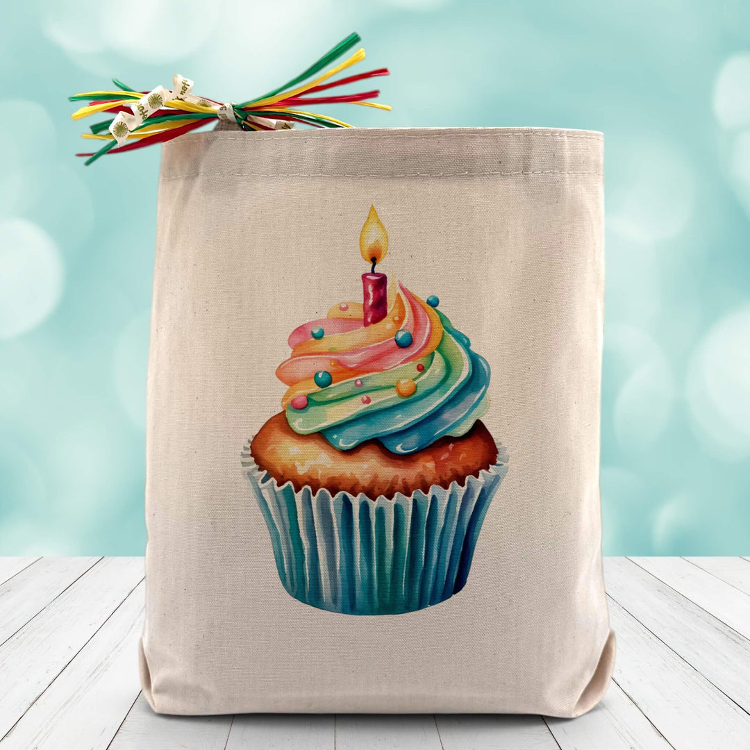 A Colorful Cupcake with sprinkles and a lit candle on top, on a canvas tote bag placed on wooden table and sparkly blue background.