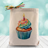 A Colorful Cupcake with sprinkles and a lit candle on top, on a canvas tote bag placed on wooden table and sparkly blue background.