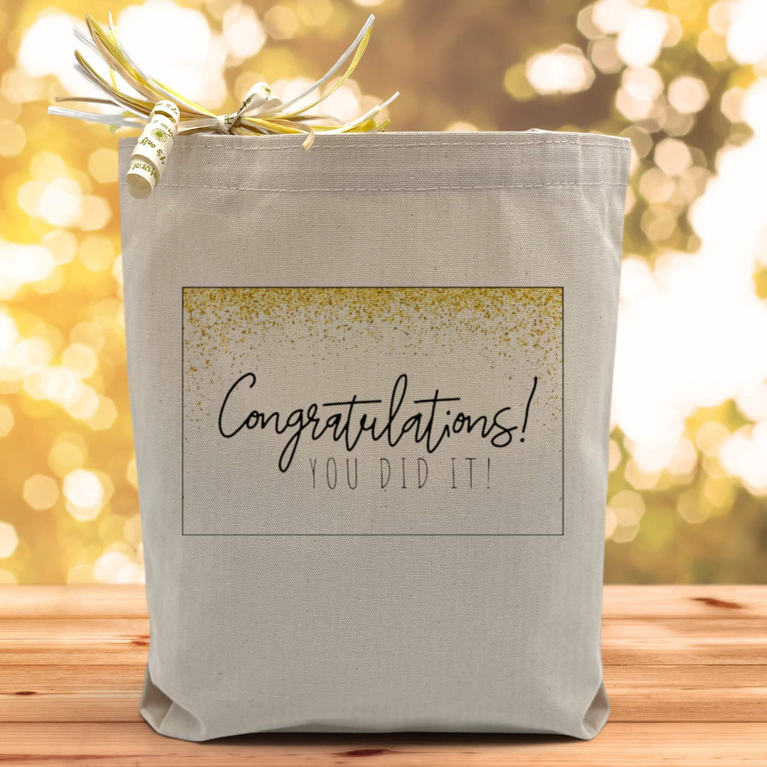 A glittery congratulations you did it text on a canvas tote bag placed on a wooden table with a sparkly background.
