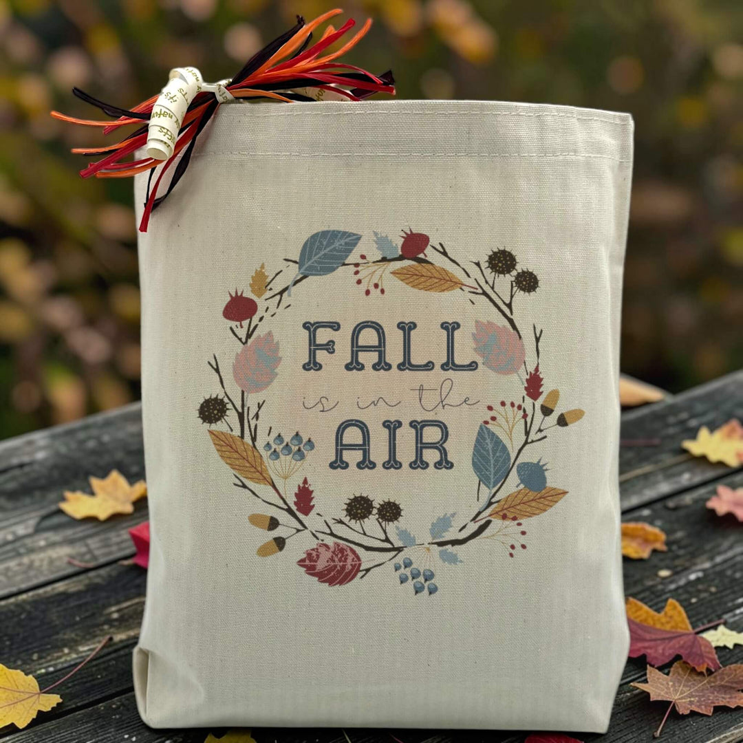 A fall is in the air text with a assortment of leaves surrounding the text on a canvas tote bag, placed on a wooden table with fall background 