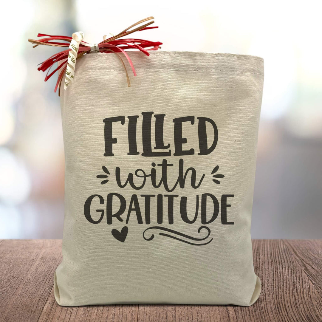 A Filled with gratitude all black text with accents and a black heart on a canvas tote bag, placed on a wooden table with a blurred grey background. 