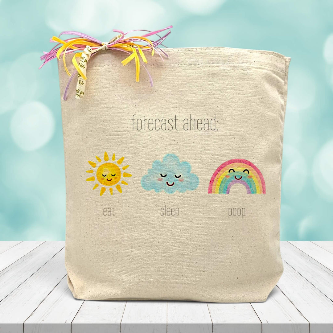 A Forecast ahead baby inspired image, with a sun, cloud, and rainbow saying eat, sleep, poop. Printed on canvas tote bag placed on wooden table with blue blurred background 