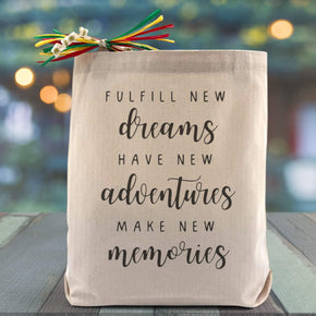 A all black text saying fulfill new dreams, have new adventures, make new memories printed on a canvas tote bag placed on a wooden table with a light filled background.