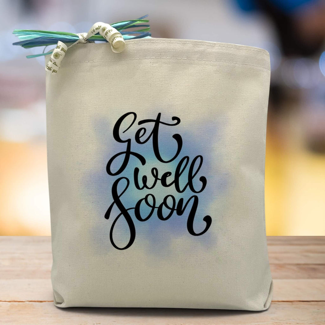 A get well soon black text with a blue airbrushed background printed on a canvas tote bag, placed on a wooden table.