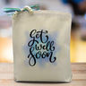 A get well soon black text with a blue airbrushed background printed on a canvas tote bag, placed on a wooden table.