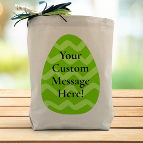 A Green easter egg with a chance to add a custom message in the middle printed on a canvas tote bag, placed on a wooden table and a leafy background.