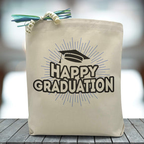 A text saying happy graduation in all black with a graduation cap printed on a canvas tote bag, set on wooden table with a grey blurred background.