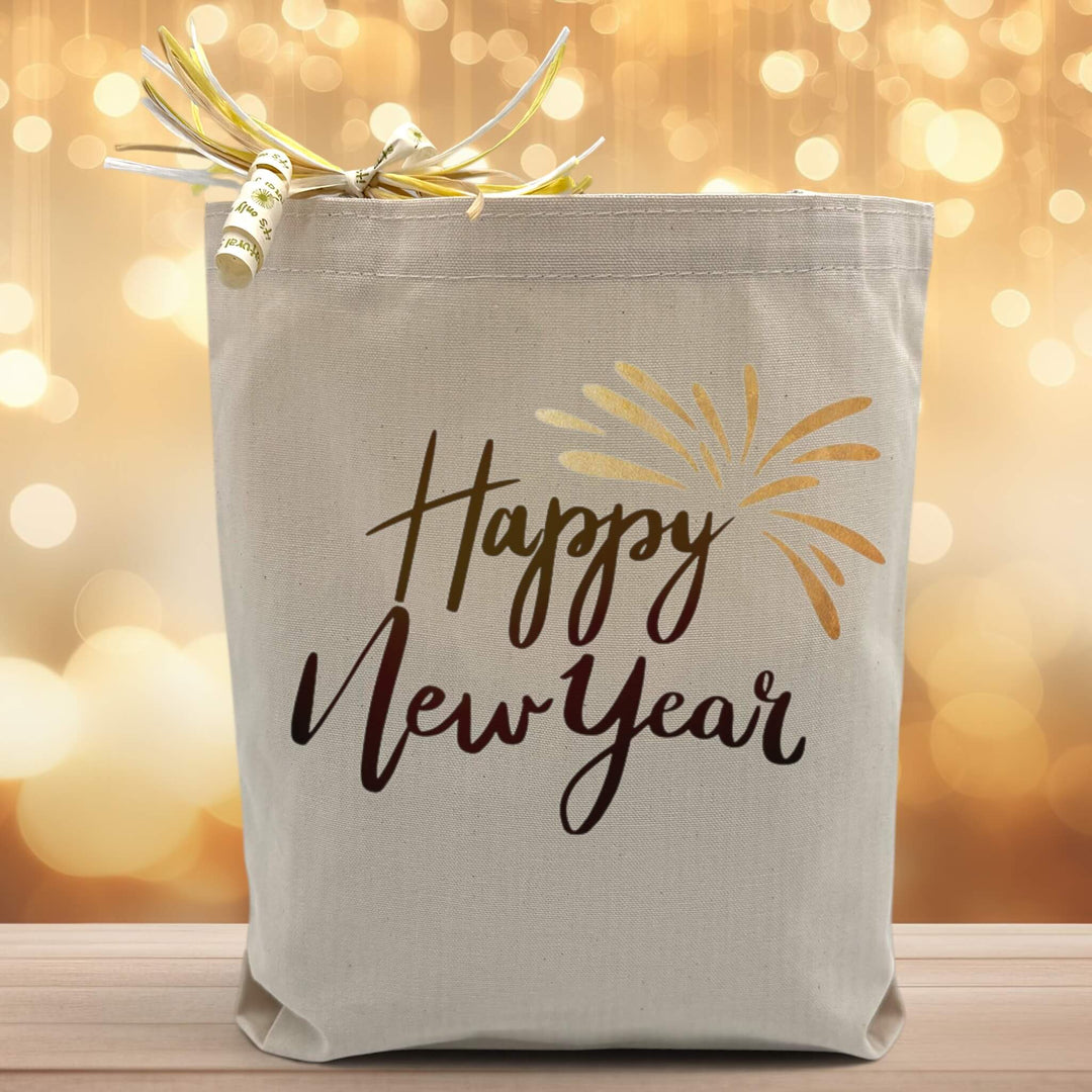 A black text saying happy new year with a gold firework accent printed on a canvas tote bag, set on a wooden table with a sparkly background.