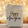 A black text saying happy new year with a gold firework accent printed on a canvas tote bag, set on a wooden table with a sparkly background.