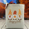 Three gnomes holding pumpkins with a happy fall text below printed on a canvas tote bag, set on a wooden table with a fall background.