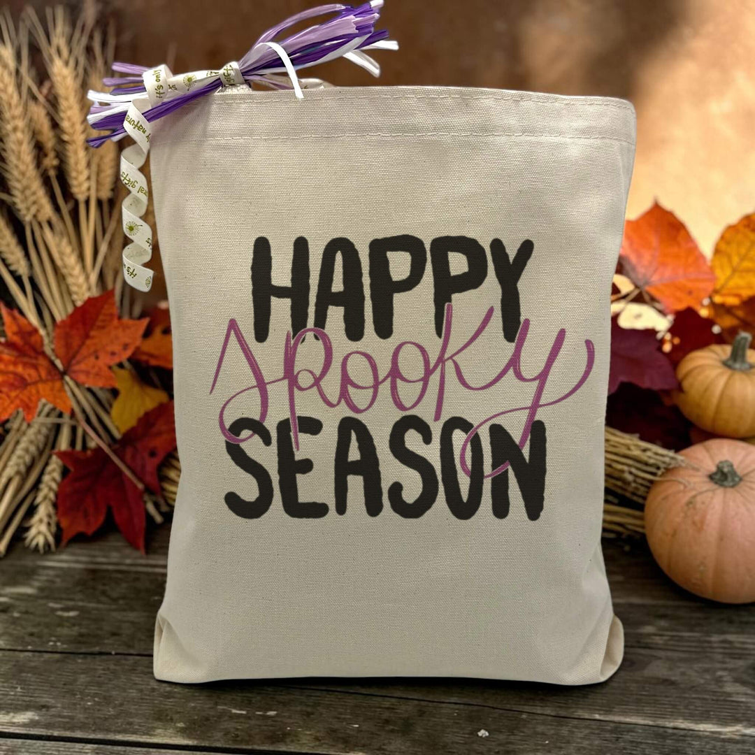 A text saying happy spooky season with only the word spooky being purple printed on a canvas tote bag, set on a wooden table with a fall inspired background.