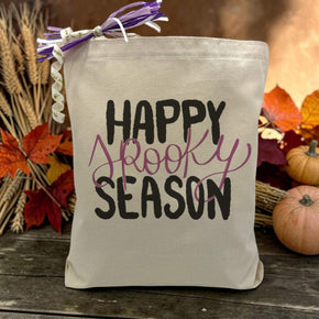 A text saying happy spooky season with only the word spooky being purple printed on a canvas tote bag, set on a wooden table with a fall inspired background.