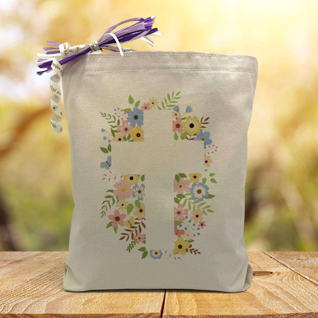 A Easter inspired cross with a floral background printed on a canvas tote bag  set on a wooden table with a light filled forest background.