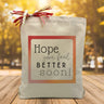 A hope you feel better soon black text with a orange, pink and yellow outline printed on a canvas tote bag set on a wooden table with a light filled forest background.