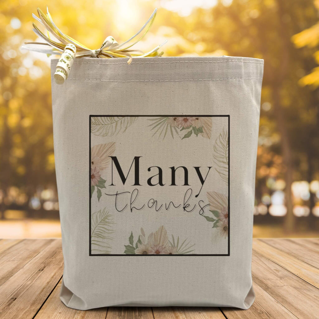 A floral image with the text many thanks placed in the middle printed on a canvas tote bag, set on a wooden table with a woodsy background.