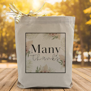 A floral image with the text many thanks placed in the middle printed on a canvas tote bag, set on a wooden table with a woodsy background.