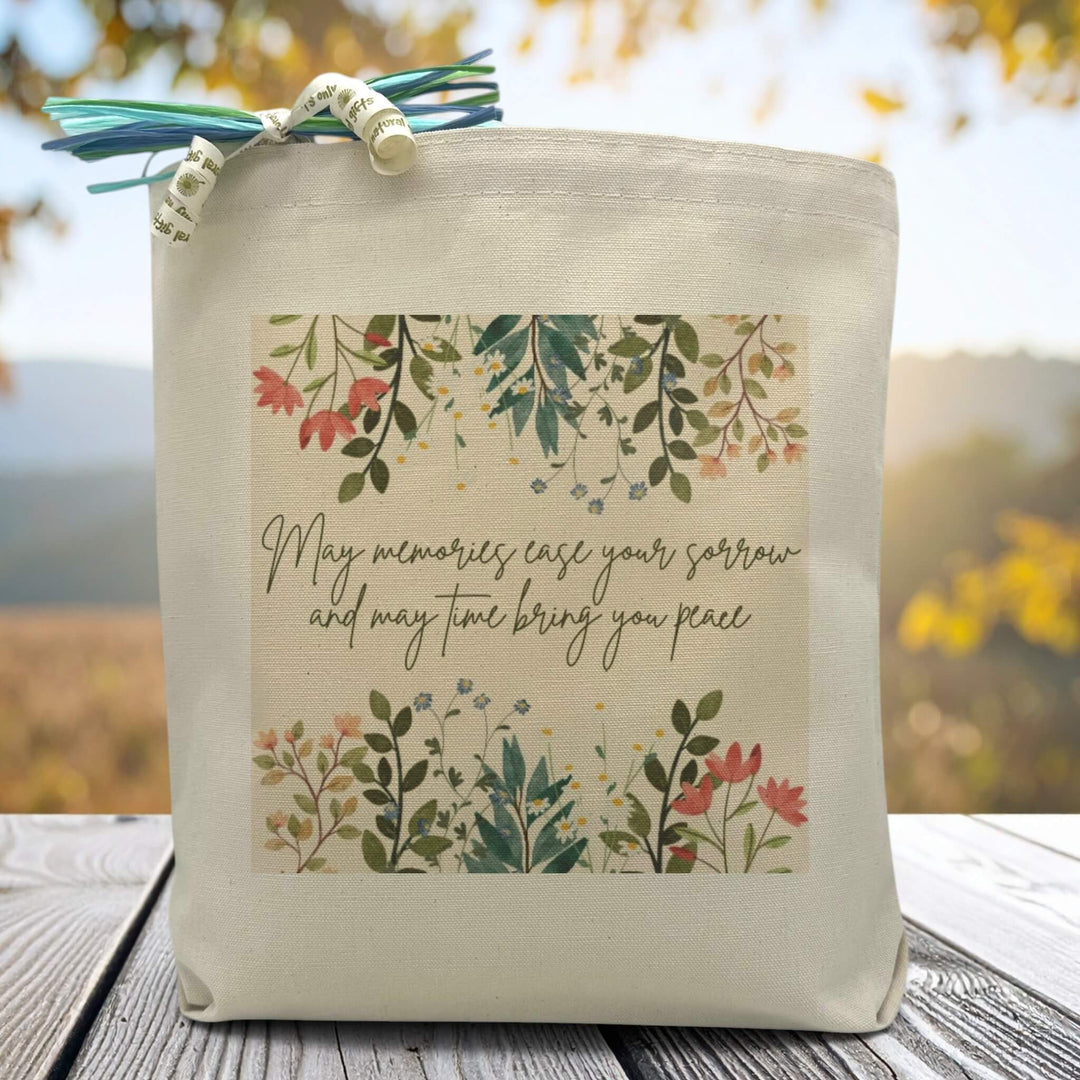 A floral background with the text may memories ease your sorrow and may time bring you peace printed on a canvas tote bag set on a wooden table.