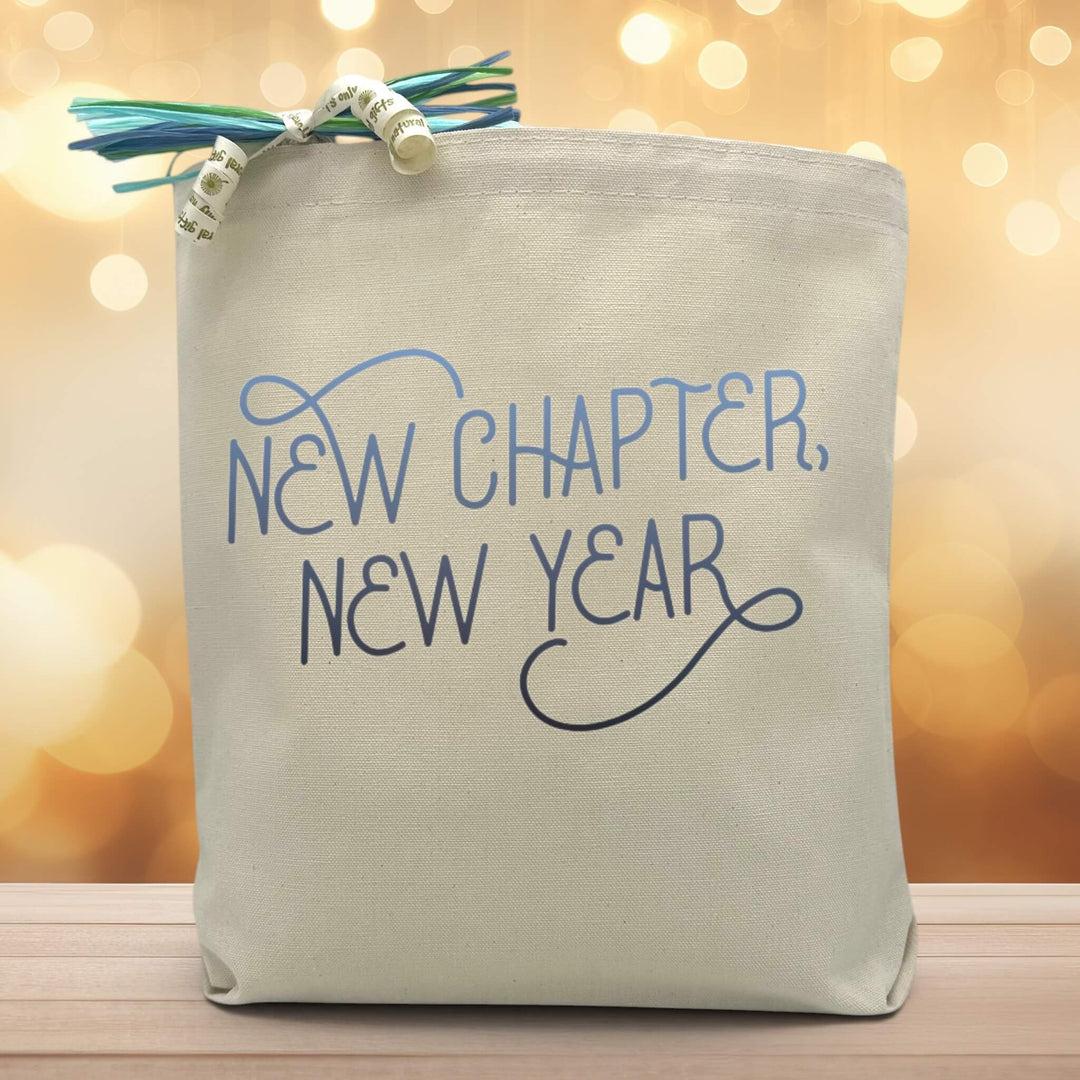 a blue text saying new chapter new year printed on a canvas tote bag, set on a wooden table with a warm light background.