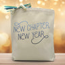 a blue text saying new chapter new year printed on a canvas tote bag, set on a wooden table with a warm light background.