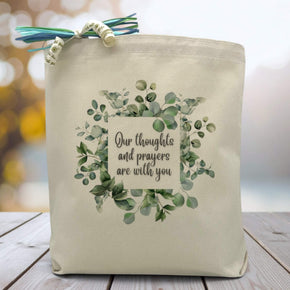 a leafy background with the text our thoughts and prayers are with you printed on a canvas tote bag set on a wooden table 