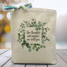 a leafy background with the text our thoughts and prayers are with you printed on a canvas tote bag set on a wooden table 