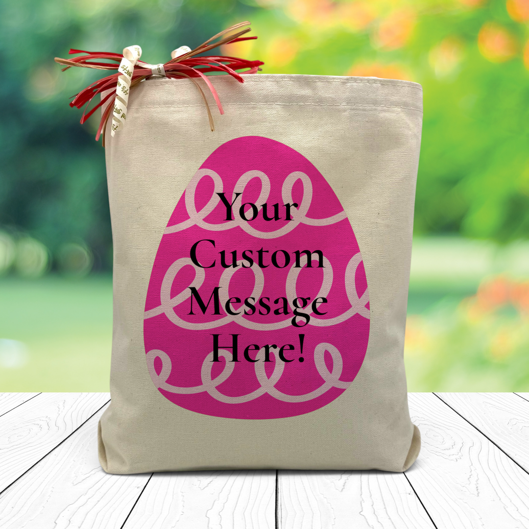 a pink easter egg with the chance to add a custom message in the middle printed on a canvas tote bag set on a wooden table with a leafy background.