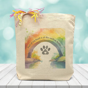 A rainbow water colored background with a text saying part of my heart is at the rainbow bridge with a paw print underneath printed on a canvas tote bag.