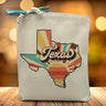 A retro styled sunrise in the shape of the Texas State with the word Texas placed in the middle printed on a canvas tote bag placed on a wooden table 