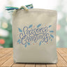 A blue text saying season's greetings with wintery accents around the text printed on a canvas tote bag placed on a wooden table with a Christmas background.