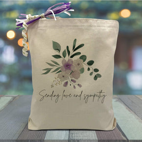  purple flowers with eucalyptus and green leaves surrounding it with a black text saying sending love and sympathy underneath printed on a canvas tote bag.