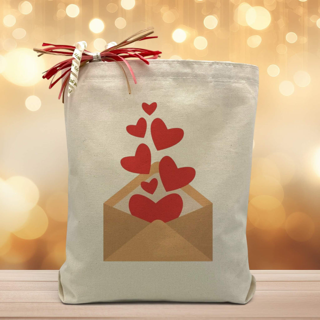 a envelope with hearts floating out of it indicating that you are sending love printed on a canvas tote bag placed on a wooden table with a light filled background. 