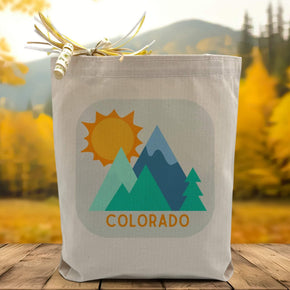  Mountains, trees and the sun with a orange colorado text beneath printed on a canvas tote bag set on a wooden table with a tree filled background.