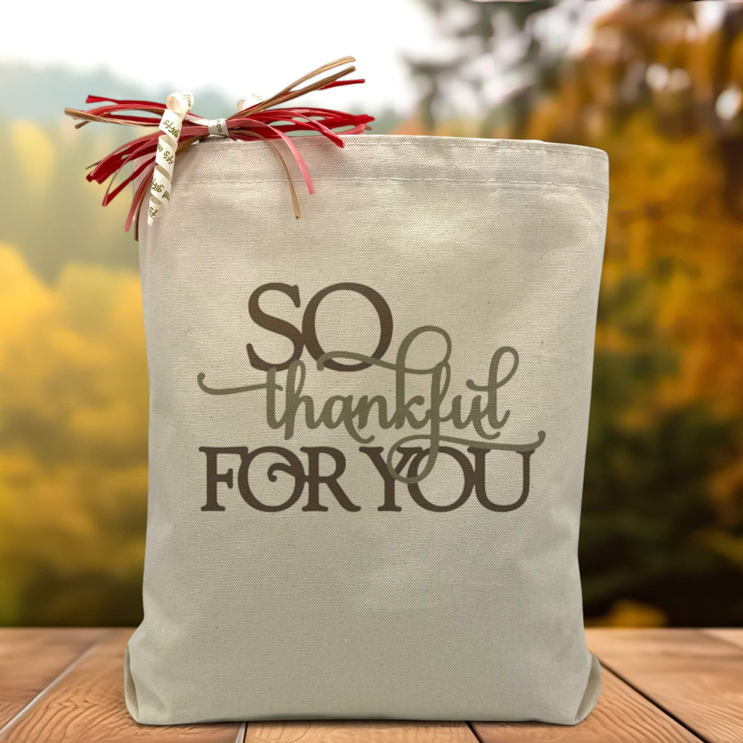 A brown and army green text saying so thankful for you printed on a canvas tote bag set on a wooden table with a tree filled background. 