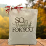 A brown and army green text saying so thankful for you printed on a canvas tote bag set on a wooden table with a tree filled background. 